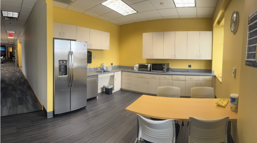An office kitchen. There is a wall of cabinets and counter space with a microwave and a toaster over. The adjacent wall has cabinets, a sink and dishwasher, and a refrigerator. There is a table and four chairs by the opposite wall.